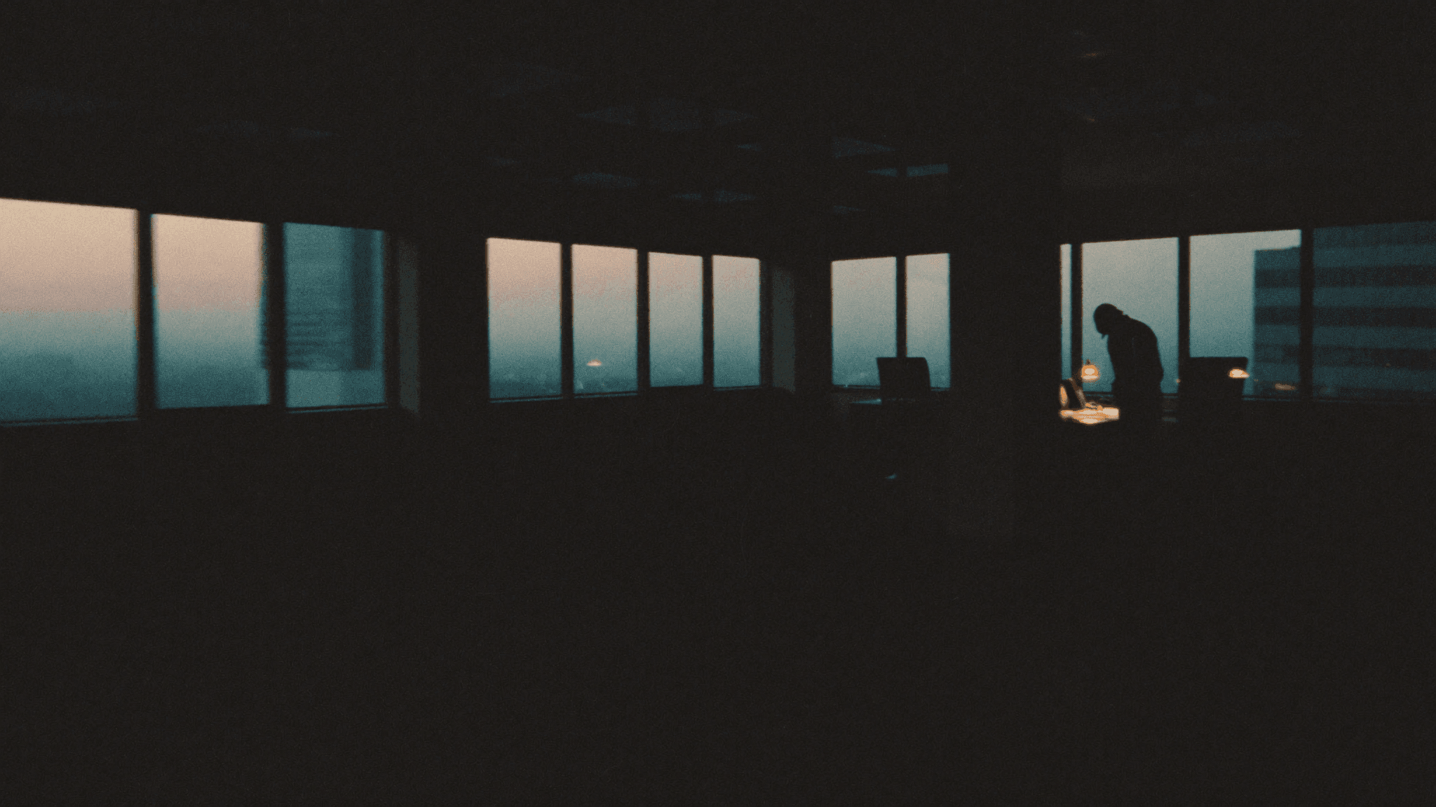 A lone figure works at a lamplit desk in a dark office before sunrise, city haze beyond floor-to-ceiling windows